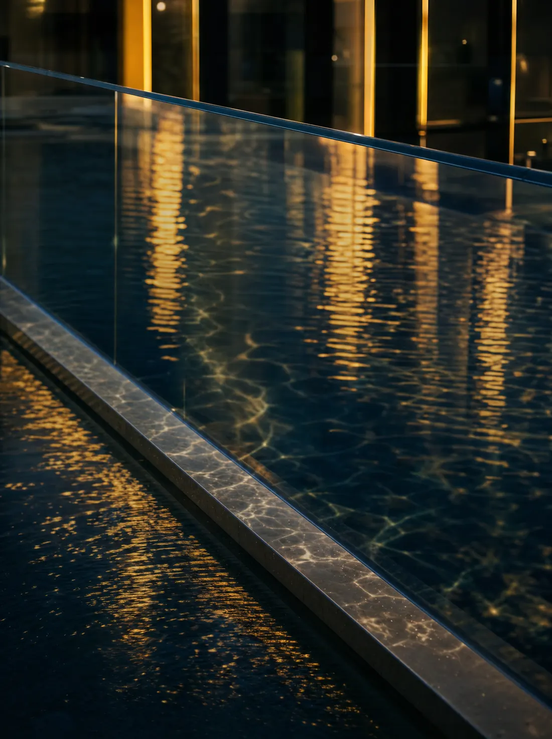 The edge of a frameless glass balustrade above a dark pool reflecting warm lights.